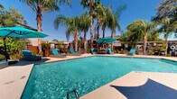 Located at 4171 E 24th St in Tucson, Arizona, this single-level outdoor space centers around a generously sized swimming pool with crystal-clear, blue-green water. The pool area is thoughtfully designed for relaxation and recreation, offering ample deck space surrounding the water's edge. Teal-cushioned lounge chairs and rattan-style chaise lounges are arranged along the pool deck, complemented by large teal shade umbrellas that provide relief from the Arizona sun. The atmosphere is defined by a striking canopy of tall, mature palm trees that frame the space, lending a resort-like ambiance characteristic of the Sonoran Desert region. Potted plants and greenery are integrated throughout the deck, adding texture and color to the hardscape. A low-profile stucco building borders one side of the pool area, featuring large windows and direct access to the outdoor space, with additional seating and dining furniture positioned nearby. A decorative water feature adds a lively element to the pool's surface. The overall setting combines the warmth of a desert climate with a lush, tropical aesthetic, creating an inviting and vibrant outdoor retreat well-suited to the sunny Tucson environment.