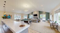 Located at 45 Fraser Blvd in Gaylord, Michigan, this single-floor community clubhouse offers a well-appointed, open-concept gathering space designed for both relaxation and recreation. The layout seamlessly integrates a kitchen, dining area, and living room into one cohesive environment.

The kitchen features a generous white quartz waterfall-edge island with warm wood bar stools, complemented by sleek gray cabinetry, a stainless steel refrigerator, and a wall-mounted television set against a bold teal accent wall. Vaulted ceilings with recessed lighting and modern pendant fixtures add to the airy, welcoming ambiance throughout.

The living area centers around a contemporary linear fireplace framed in light tile, with a wall-mounted TV above and comfortable sectional seating nearby. A pool table serves as a central recreational feature, adding a social and lively dimension to the space. Hardwood-style flooring transitions to plush carpeting in the lounge zones, grounding each area distinctly.

Large windows and glass sliding doors flood the interior with natural light while offering direct views of the outdoor pool and surrounding greenery. A dedicated dining nook with bistro-style seating and a small workspace area round out the functionality of this versatile, community-focused space.