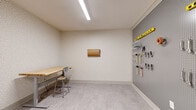 This single-floor workshop space is thoughtfully designed to support hands-on work in a clean and organized setting. The room features light vinyl plank flooring throughout and walls finished with a subtle, textured wallcovering that lends a polished look uncommon in traditional workshop environments. Overhead, recessed linear LED lighting ensures bright, even illumination across the entire workspace.

The focal point of the room is a large floor-to-ceiling pegboard wall finished in gray, neatly outfitted with a comprehensive collection of hand tools — including hammers, mallets, pliers, wrenches, screwdrivers, clamps, and levels — all visibly organized and within easy reach. A height-adjustable workbench with a solid wood top and an industrial metal stool provides a dedicated surface for projects of varying nature.

A wall-mounted paper roll dispenser adds practical functionality for wrapping or protecting surfaces. A compact utility area near the entrance includes warm wood cabinetry and a basin sink, supporting cleanup and prep tasks. The space is accessed through a glass-paneled door that opens to a corridor, connecting this workshop to the broader facility. Together, these elements create a highly functional, well-appointed workspace suited for a range of craft, repair, or DIY activities.