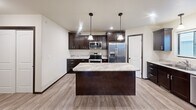 Located in Box Elder, South Dakota, this single-floor, 2-bedroom, 2-bathroom home offers a thoughtfully designed layout with modern finishes throughout. The open-concept living, dining, and kitchen area forms the heart of the home, anchored by light wood-look flooring and a ceiling fan that enhances the bright, airy atmosphere. The kitchen is a standout feature, boasting rich dark cabinetry, granite-style countertops, stainless steel appliances including a range, microwave, French door refrigerator, and dishwasher, as well as a central island with pendant lighting above — ideal for both meal prep and casual entertaining. A pantry adds convenient additional storage. Both bedrooms are comfortably sized, featuring plush carpet, ceiling fans, and natural light from windows with blinds. Multiple closets throughout provide ample storage. The two bathrooms are finished with dark wood vanities, granite countertops, and full tub-shower combinations with wood-look flooring. Additional practical spaces include a dedicated laundry room, utility room, hallway, and a balcony for outdoor enjoyment. The consistent use of dark trim, neutral tones, and quality materials lends a cohesive, contemporary feel to the entire home.