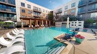 Located at 1100 S Cesar Chavez Blvd in Dallas, TX, this meticulously designed outdoor amenity space serves as the central courtyard of a modern multi-story residential community. The exterior architecture combines brick masonry with contemporary metal paneling and large-format windows, while private balconies overlook the grounds on multiple levels above.

At the heart of the courtyard is a generously sized resort-style swimming pool with crystal-clear blue water, surrounded by a spacious concrete deck furnished with lounge chairs and umbrellas for sunbathing. A pergola structure anchors one end of the pool deck, providing shaded seating and an architectural focal point. Tiered water features with stone-filled channels add a tranquil, decorative element near the pool's edge.

The surrounding landscape is thoughtfully designed with mature trees, manicured shrubs, ornamental grasses, and colorful plantings that provide greenery and privacy throughout. A central lawn area offers open green space, complemented by paved walkways, decorative lampposts, and outdoor seating areas with tables and chairs. Iron fencing and low brick walls define pathways that wind through the courtyard, connecting various amenity zones. The overall ambiance is open, bright, and well-maintained, creating an inviting outdoor retreat within an urban Dallas setting.