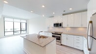 This single-floor residence presents a contemporary, open-concept layout bathed in natural light. The kitchen serves as a central focal point, featuring high-gloss white cabinetry extending to the ceiling, a subway tile backsplash, and granite countertops throughout. A generously sized center island with an integrated sink and matching granite surface provides both additional prep space and a natural gathering point. Stainless steel appliances, including a built-in microwave, electric range, and refrigerator, complete the modern kitchen package. Recessed lighting illuminates the space efficiently.

Light-toned hardwood flooring flows seamlessly from the kitchen into the open living area, which benefits from floor-to-ceiling windows framing expansive city views. The bedroom, accessible from the living space, also features large windows allowing abundant natural light.

The bathroom is finished with warm-toned ceramic tile flooring and includes a white vanity with granite countertop, a toilet, and a glass-enclosed walk-in shower accented with a decorative mosaic tile inlay. The overall design palette—white walls, light wood floors, and neutral stone surfaces—creates a cohesive, clean aesthetic throughout the unit.