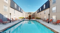 Located at 2400 Westheimer Rd in Houston's vibrant Montrose neighborhood, this single-floor property is part of a multi-building complex that combines classic brick architecture with a dark mansard-style roofline, creating a distinctive and cohesive aesthetic. The complex is centered around a generously sized outdoor swimming pool, framed by a spacious sun deck furnished with lounge chairs and shaded seating beneath red umbrellas — an inviting space for relaxation and recreation. The buildings surrounding the courtyard feature large, multi-pane windows that allow abundant natural light throughout, while the exterior brick facades lend a timeless, traditional character. Covered exterior walkways with arched brick columns and black wrought-iron railings provide sheltered access between units, adding both architectural charm and practical convenience. Sliding glass doors open directly onto these walkways, connecting interior living spaces to the outdoor corridor and courtyard beyond. The overall ambiance of the property is one of a well-established, community-oriented setting, with lush potted plantings and landscaped beds softening the hardscape around the pool deck. Its prime location along Westheimer Rd places residents within easy reach of Houston's best dining, shopping, and entertainment destinations.