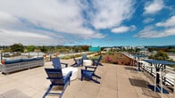 Located at 555 Pacific Ave in Santa Cruz, California, this rooftop space offers a distinctive outdoor environment designed with sustainability and usability in mind. The rooftop is defined by expansive green roof plantings featuring a diverse selection of drought-tolerant succulents, ornamental grasses, flowering shrubs, and ground covers arranged across wide landscaped beds on either side of a central concrete walkway. The walkway runs the length of the rooftop, connecting multiple outdoor seating areas furnished with tables, chairs, and lounge seating suited for both casual gatherings and quiet relaxation. A vibrant teal stairwell enclosure serves as a notable architectural accent and provides rooftop access. The perimeter is lined with a modern horizontal-rail railing system that ensures safety while preserving unobstructed sightlines. Panoramic views extend in all directions, encompassing the Santa Cruz cityscape, neighboring rooftops, tree-lined residential streets, and the rolling hills in the distance. The open sky overhead and abundant natural light enhance the overall ambiance, making this rooftop a welcoming outdoor retreat. The thoughtfully designed green roof not only adds visual character but also reflects an environmentally conscious approach to urban design.