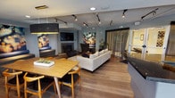 The Club Room at 1800 S Jackson Street in Seattle, Washington, is a thoughtfully designed communal space that offers residents a sophisticated and welcoming environment for socializing and relaxation. The single-floor layout flows seamlessly from a dining area to a comfortable lounge, creating a versatile setting for a variety of gatherings. Warm hardwood-style flooring runs throughout, tying the space together with a cohesive, contemporary aesthetic. The lounge area is anchored by a sleek fireplace framed by a wall-mounted television, providing a cozy focal point surrounded by plush seating, including a generous sectional sofa and accent chairs. Overhead, adjustable track lighting and pendant fixtures allow for customizable illumination, while recessed ceiling lights add to the layered lighting scheme. A dining area with a natural wood table and mid-century modern chairs offers an inviting spot for shared meals or casual meetups. Large windows dressed with sheer drapes allow natural light to filter into the space, with access to an adjacent area visible through the glass. Curated artwork and potted greenery add personality and warmth, complementing the cool-toned walls. This Club Room delivers a polished yet comfortable atmosphere, ideal for community events and everyday use.