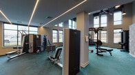 This two-floor fitness facility is thoughtfully designed to accommodate a wide variety of training styles and fitness goals. The main floor is expansive and bathed in natural light streaming through large black-framed windows that offer views of the surrounding landscape. The space is grounded by a distinctive teal patterned carpet and illuminated by sleek linear LED lighting recessed into the ceiling, lending the environment a modern, polished aesthetic.

The main workout floor is well-equipped with a comprehensive selection of strength and cardio machines, including treadmills, ellipticals, stair climbers, and multi-station cable machines. A dedicated free weights section features a full rack of dumbbells alongside medicine balls and benches. A functional training zone includes a power rack, gymnastic rings, battle ropes, and a heavy bag, catering to more dynamic and athletic training styles. Floor-to-ceiling mirrors line key walls, enhancing the sense of space throughout.

An interior staircase — accented by a decorative patterned wall — connects to the second level, which provides additional cardio equipment and overlooks the main floor below. A separate studio room with light hardwood flooring, deep navy walls, and mirrored walls houses Pilates reformers and serves as a dedicated space for focused, low-impact training. Together, both floors offer a complete and well-appointed fitness experience.