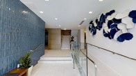 This contemporary lobby makes a striking first impression through a thoughtful combination of modern design elements and curated artistic touches. The space is anchored by a textured deep-blue accent wall that adds visual depth and character, contrasting beautifully with the light, neutral tones found throughout the rest of the interior. Large-format white tile flooring reflects the abundant recessed lighting overhead, contributing to a bright and open atmosphere. A central staircase with clean white steps and polished gold metal railings serves as a focal point, guiding visitors upward with understated elegance. Sleek glass panel railings with stainless steel hardware line the walkways, reinforcing the space's modern aesthetic while maintaining an open sightline throughout. A standout sculptural installation of blue and white abstract forms adorns one wall, infusing the lobby with artistic energy and a distinct sense of identity. Greenery in the form of potted plants adds a natural, welcoming touch. The overall ambiance is polished and professional, balancing clean architectural lines with warm design accents to create a lobby that is both functional and visually impressive.