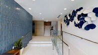 This contemporary lobby makes a striking first impression through a thoughtful combination of modern design elements and curated artistic touches. The space is anchored by a textured deep-blue accent wall that adds visual depth and character, contrasting beautifully with the light, neutral tones found throughout the rest of the interior. Large-format white tile flooring reflects the abundant recessed lighting overhead, contributing to a bright and open atmosphere. A central staircase with clean white steps and polished gold metal railings serves as a focal point, guiding visitors upward with understated elegance. Sleek glass panel railings with stainless steel hardware line the walkways, reinforcing the space's modern aesthetic while maintaining an open sightline throughout. A standout sculptural installation of blue and white abstract forms adorns one wall, infusing the lobby with artistic energy and a distinct sense of identity. Greenery in the form of potted plants adds a natural, welcoming touch. The overall ambiance is polished and professional, balancing clean architectural lines with warm design accents to create a lobby that is both functional and visually impressive.