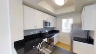 This single-story space showcases a well-appointed kitchen that combines modern aesthetics with practical design. The kitchen is outfitted with crisp white shaker-style cabinetry that extends to the upper walls, providing generous storage throughout. Dark granite countertops create a striking contrast against the white cabinets, lending a polished, contemporary feel to the space.

Stainless steel appliances, including a range with an electric cooktop, built-in microwave, and a full-sized refrigerator, are seamlessly integrated into the layout. A stainless steel sink with a modern arc faucet is positioned at the countertop, complementing the overall finish palette.

The flooring transitions to warm-toned hardwood, adding a natural element that balances the kitchen's modern touches. Natural light filters in through a window fitted with vertical blinds, brightening the space alongside a flush-mount ceiling light fixture. The tray ceiling detail above adds subtle architectural interest, contributing to the clean, structured character of the kitchen's design.