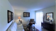 This well-appointed single-floor residence offers a warm and welcoming atmosphere anchored by a thoughtfully designed living area. The living room serves as the heart of the space, featuring plush white seating, a sleek round coffee table, and a cozy accent chair, all arranged atop a richly patterned area rug. Smooth, light-colored walls provide a clean backdrop that enhances the sense of openness throughout the room. A framed piece of wall art adds a touch of personality, while a classic table lamp contributes soft, ambient lighting to complement the room's overall warmth. Natural light flows generously into the space through a large window dressed with vertical blinds, offering a bright and airy feel. The neutral carpeted flooring ties the room together seamlessly, creating a cohesive and comfortable environment. The overall ambiance is calm and functional, with a layout that balances open space and purposeful furnishing placement, making it an ideal setting for both relaxing and entertaining.