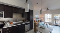 Located at 450 10th in San Diego, this model unit presents a thoughtfully designed single-floor layout that balances contemporary style with functional living. The open-concept kitchen is a standout feature, showcasing rich dark cabinetry that extends floor to ceiling, a tiled backsplash, light quartz countertops, and a full suite of stainless steel appliances including a refrigerator, range, microwave, and dishwasher. A double-basin sink and ample storage make this kitchen as practical as it is attractive. The kitchen flows seamlessly into the dining and living areas, where warm wood-look flooring runs underfoot and pendant lighting adds a modern touch overhead. The living room is bright and welcoming, with large windows and a glass door opening to a private balcony that frames views of the surrounding San Diego skyline. A ceiling fan adds comfort to this airy space. The bedroom is a calm retreat, featuring wood-look flooring, a generous window with urban views, and neutral tones throughout. The bathroom is clean and modern, with dark vanity cabinetry, a white countertop, tile flooring, and a shower-tub combination. Throughout the unit, the design palette of dark accents against light walls and surfaces creates a cohesive, contemporary aesthetic.