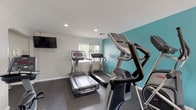 This fitness center is a thoughtfully designed, single-floor facility offering a comprehensive workout environment for users of all fitness levels. The space is divided into distinct functional zones, seamlessly connected to encourage a full-body training experience.

The cardio area is outfitted with multiple treadmills and elliptical machines from recognized equipment brands, positioned to take advantage of natural light streaming in through several windows. A wall-mounted flat-screen television provides entertainment during workouts, while a vibrant teal accent wall adds a bold, energizing visual element against the otherwise neutral, light-gray walls.

The strength training zone features a selection of selectorized weight machines, including a cable/functional trainer and a seated rowing machine, catering to a range of resistance exercises. A large wall mirror spans one side of the room, enhancing the sense of space and allowing users to monitor their form during exercise. The mirror is accompanied by a ballet-style barre rail, suggesting the space may also support stretching or light movement routines.

Throughout the facility, durable dark rubber flooring provides safety and sound absorption. Recessed lighting and HVAC vents in the ceiling ensure consistent comfort, while multiple windows offer views of the surrounding greenery. The overall ambiance is clean, functional, and well-maintained.