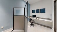 Located at 5961 SW 68th St in South Miami, FL, this compact yet thoughtfully designed office workspace offers a clean and professional environment well-suited for focused work. The space is organized around a continuous marble-topped built-in desk that runs along one wall, providing ample surface area for computers, printing equipment, and other office essentials. A sleek all-in-one desktop computer and multifunction printer are integrated into the workstation, complemented by ergonomic mesh task chairs that prioritize comfort during extended use. The interior palette features soft blue-gray walls that lend a calm, contemporary feel to the room. Bold blue abstract artwork displayed along the walls adds a touch of creative character while reinforcing the cohesive color scheme. Light-toned tile flooring enhances the brightness of the space and is easy to maintain. A modern glass-panel partition with a dark metal frame defines the workspace boundary, offering both openness and a degree of visual separation. Entry to the space is provided through a full-height glass sliding door with a dark-framed surround, contributing to the polished, professional aesthetic. Recessed ceiling lighting ensures consistent, even illumination throughout. Overall, this office workspace balances functional design with refined finishes, creating an efficient and visually appealing environment for professional use.