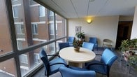 This single-floor space serves as a comfortable and functional waiting or meeting area, thoughtfully designed to promote a calm and welcoming atmosphere. The most striking architectural feature is the expansive floor-to-ceiling windows that line one wall, flooding the interior with natural light and offering views of the surrounding brick building exterior. The tile flooring grounds the space with a clean, professional aesthetic, while the drop ceiling with warm wall-mounted lighting adds a soft, inviting glow. At the center of the room sits a round table surrounded by cushioned blue chairs, creating an approachable setting for conversation or casual meetings. Additional blue seating is arranged along the perimeter, offering flexible accommodation for multiple occupants. Potted plants placed on the table and in the corner introduce a touch of greenery, enhancing the overall sense of comfort and warmth. Decorative wall art adds a subtle personal touch to the otherwise neutral, cream-toned walls. The layout is open and uncluttered, making efficient use of the available space while maintaining a relaxed, accessible environment suitable for a variety of professional or institutional settings.