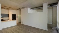 This single-floor space showcases a compelling industrial-modern aesthetic that blends raw architectural elements with clean, contemporary finishes. The exposed concrete ceiling immediately draws the eye upward, lending the interior a loft-like character and an open, airy feel. A prominent cylindrical metal duct runs horizontally along the ceiling, adding to the industrial charm while serving a functional purpose.

Track lighting mounted to the ceiling provides flexible, directional illumination across the main living area, complementing the natural brightness of the space. The floors are finished with wide-plank wood-look material in a warm gray-brown tone, offering both durability and visual warmth that contrasts beautifully with the raw overhead surfaces.

The main living area flows seamlessly toward a kitchen section, where dark cabinetry and a light-colored countertop create a striking contrast. A hallway visible beyond the kitchen leads to additional areas of the unit, suggesting a thoughtfully laid-out floor plan. White walls throughout keep the space feeling fresh and versatile, providing a neutral canvas ready for personalization. Overall, the space strikes a confident balance between urban industrial character and modern livability.