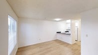 This single-floor space features an open-concept design that seamlessly connects the living area to the kitchen. The living space is finished with light hardwood flooring that adds warmth to the neutral white walls, creating a clean and bright environment. A textured ceiling with flush-mount lighting provides even illumination throughout the room.

The kitchen is accessible via a pass-through counter with a dark contrasting countertop, which also functions as a breakfast bar. White cabinetry lines the kitchen walls, complementing the white appliances, including a refrigerator and stove. A stainless steel sink sits beneath the countertop opening, adding a functional and modern touch. The kitchen's layout is compact yet efficient, offering practical workspace.

The overall ambiance of the space is minimal and functional, with a consistent use of white tones and light wood flooring tying the areas together. The open sightlines between the living and kitchen areas enhance the sense of spaciousness, making the unit feel airy and easy to navigate.