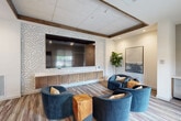 Located at 1700 Katy Fort Bend Road in Katy, Texas, this contemporary lounge is a single-room living space designed with modern sophistication. The room's most striking feature is a bold circuit board-patterned accent wall that frames a large flat-screen television, lending a tech-forward aesthetic to the space. Beneath the screen, a built-in wooden media console provides sleek, functional storage. The seating area is anchored by a set of curved, deep-blue velvet lounge chairs paired with natural wood cube side tables, creating a comfortable and visually cohesive gathering space. Underfoot, a multicolored striped carpet adds warmth and texture, complementing the wood-toned flooring throughout. Exposed wooden ceiling beams add an architectural dimension to the tray ceiling above, enhancing the room's character. Large black-framed windows along one wall flood the interior with natural light and offer views of the outdoor patio area. A compact wet bar or kitchenette nook with cabinetry and a stainless steel appliance is tucked to one side, adding convenience. Abstract wall art and a lush fiddle-leaf fig plant complete the refined, welcoming ambiance of this well-appointed lounge.