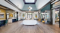 This thoughtfully designed single-floor amenity space offers a generously scaled, open layout that blends modern aesthetics with inviting functionality. The entrance greets visitors with warm wood-look flooring, decorative wallcoverings in soft gold tones, and glass-paneled double doors that flood the foyer with natural light and outdoor views.

The main lounge area commands attention with a dramatic vaulted ceiling painted in a calming sky blue, anchored by a sculptural globe pendant chandelier. A curved white sofa, layered area rugs, and vibrant artwork create an energizing yet comfortable atmosphere. Large floor-to-ceiling windows and glass doors provide direct sightlines to an outdoor pool area, enhancing the sense of openness throughout.

Adjacent to the lounge is a versatile gathering zone featuring a round conference table, upholstered task chairs, a decorative fireplace with a wood mantel, and a wall-mounted television framed by intricate patterned tile—ideal for both casual meetings and social events.

The kitchenette impresses with deep navy cabinetry, quartz countertops, and bold geometric-patterned tile backsplash, offering a functional yet design-forward refreshment area. A dedicated billiards room rounds out the space, showcasing soaring teal-painted walls, oversized windows with transom details, and stylish pendant lighting above a full-sized pool table.