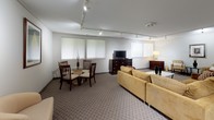 Located at 3200 S Adams Rd in Auburn Hills, Michigan, this well-appointed single-floor suite offers a comfortable and functional layout designed for both relaxation and productivity. The generously sized main living area is finished with patterned wall-to-wall carpeting and features track lighting that runs the length of the ceiling, providing flexible, adjustable illumination throughout the space. Multiple windows fitted with white horizontal blinds allow for natural light while maintaining privacy. The living area is furnished with a sectional sofa, accent chairs, glass-top coffee tables, and dark wood dresser units that house a flat-screen television, creating a cohesive and welcoming atmosphere. A dedicated dining area with a round glass table and wooden chairs sits adjacent to the main seating zone, offering a space for meals or casual meetings. A dark wood armoire and a framed wall piece add refined touches to the decor. Crown molding along the upper walls of the living area adds an architectural detail that elevates the overall aesthetic. The kitchen is fully equipped with white cabinetry, a refrigerator, electric range, microwave, and a sink set into a peninsula countertop — offering full meal preparation capability. Light tile flooring in the kitchen contrasts neatly with the carpeted living spaces, clearly defining each functional zone.
