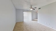 This single-floor unit offers a practical and efficient layout within a cozy footprint. The living area is finished with neutral beige carpeting and painted in a soft gray tone, creating a calm and understated atmosphere. A ceiling fan with an integrated light fixture provides both ventilation and ambient lighting, while a set of vertical blinds covers a sliding glass door that opens to the outdoors, allowing natural light to filter in. The textured popcorn ceiling adds a classic residential touch throughout the main living space. Adjacent to the living area, a dedicated dining space is highlighted by a pendant light fixture and a large window fitted with horizontal blinds, filling the area with natural light. The kitchen is positioned alongside the dining area and features white cabinetry, a tile backsplash, and standard appliances including a stovetop range and refrigerator. A pass-through or bar-height counter connects the kitchen to the adjacent space, enhancing the open feel. The bedroom is generously proportioned relative to the overall layout, also featuring a ceiling fan and carpeted flooring. Dark hardwood-style flooring appears at the transition points near the bedroom and kitchen areas, adding a subtle contrast to the soft carpeting throughout the rest of the unit.