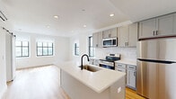Located at 933 McGee St, Unit 1104 in Kansas City, Missouri, this thoughtfully designed unit offers a clean, modern aesthetic across a single floor. Warm hardwood-style flooring runs consistently throughout the space, creating a cohesive and inviting atmosphere.

The open-concept kitchen serves as the heart of the unit, featuring gray shaker-style cabinetry, stainless steel appliances including a range, microwave, and refrigerator, and a white quartz-topped center island with an undermount sink. A subway tile backsplash and large black-framed window add character and allow natural light to fill the space. A wall-mounted mini-split HVAC unit provides efficient climate control.

The living area is bright and open, connected seamlessly to the kitchen via an open doorway. It includes a ceiling fan, a wall-mounted smart thermostat, and an additional mini-split unit, ensuring year-round comfort.

A sliding barn door leads to the private hallway, which connects to both the bathroom and walk-in closet. The bathroom features a glass-enclosed walk-in shower with tile surround and matte black hardware accents. The spacious walk-in closet is equipped with wire shelving on both sides, offering ample storage. Recessed lighting throughout the unit contributes to a clean, modern finish.