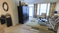 This single-floor living space offers a warm and inviting atmosphere defined by an abundance of natural light streaming through large windows dressed with plaid drapery panels. The generous window placement creates a bright, airy environment throughout the day. Soft, neutral-toned wall-to-wall carpeting anchors the space, complemented by a patterned area rug in muted earth tones — greens, tans, and grays — that adds visual depth and definition to the seating area. A bold, dark-stained armoire serves as a striking focal point against the light-colored walls, topped with decorative accessories including a vase and framed artwork, adding character to the room. Crown molding along the upper walls introduces an architectural detail that lends a refined, classic touch. A sleek, black metal-framed coffee table sits at the center, paired with a cushioned accent chair in neutral tones for a cohesive, understated aesthetic. A glass-paneled door provides access to an exterior area, further enhancing the connectivity between indoor and outdoor spaces. Overall, the room presents a well-balanced blend of functional design and tasteful décor.