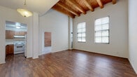 This single-floor space makes a striking architectural impression with its soaring ceilings adorned by a series of warm, exposed wood beams that span the length of the main living area. Rich hardwood flooring runs throughout the open living spaces, complementing the crisp white walls and lending a warm, inviting character to the interior. Large gridded windows flood the main areas with generous natural light, offering views of the surrounding exterior. The spacious main living area is open and airy, benefiting fully from the dramatic ceiling height and beam detailing above. The kitchen is well-appointed with warm wood cabinetry, ample counter space, an electric range, and a stainless steel sink, all set atop matching hardwood floors. The bedrooms are carpeted for comfort, each featuring tall ceilings, large windows with blinds, and closet space with bifold doors for practical storage. Two bathrooms serve the space: one with a full tub-shower combination, a wood-vanity, and tile flooring, and a second featuring a walk-in shower with a glass door, a matching wood vanity, and light tile floors. An additional area with exposed brick detailing adds a distinctive, industrial-chic touch. Overall, the space blends loft-style architectural character with comfortable, functional living areas.