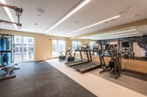 This contemporary fitness center is thoughtfully designed to accommodate a full range of workout routines within a single, open floor layout. The space is finished with a clean, neutral color palette — warm beige walls complemented by dual-tone flooring that separates the cardio zone from the strength and functional training areas.

The cardio section lines one side of the room and includes multiple treadmills and stationary bikes, each equipped with individual screens for entertainment or performance tracking. A floor-to-ceiling mirror runs along this zone, amplifying the sense of space and allowing users to monitor their form.

The strength training area features a robust power rack outfitted with battle ropes and accessory attachments, a multi-tier dumbbell rack stocked with a wide range of weights, kettlebells, medicine balls, and a cable resistance machine — offering a comprehensive setup for both compound and isolated movements. An open stretch of dark rubber flooring provides ample room for functional exercises and floor work.

Linear LED lighting runs across the ceiling, delivering bright, even illumination throughout. Large windows and a glass sliding door flood the space with natural light and offer views of the surrounding exterior. The overall ambiance is clean, professional, and energizing — well-suited for focused, high-quality training sessions.