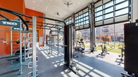 This fitness center at 3710 Governors Dr SW in Huntsville, Alabama, is a thoughtfully designed, single-story facility that combines high-end equipment with an energizing architectural aesthetic. The space is defined by dramatic floor-to-ceiling glass garage-style doors that span an entire wall, flooding the interior with abundant natural light and offering unobstructed views of a landscaped outdoor courtyard. When open, these doors seamlessly merge the indoor workout area with the outdoors, creating an inviting open-air atmosphere.

The main workout floor features rubber flooring throughout and is equipped with a comprehensive selection of TKO strength training systems, including functional rigs with pull-up bars, cable machines, and a heavy punching bag. A dedicated cardio zone houses a row of True and Inmotion treadmills, ellipticals, stationary bikes, and a Shi-Row rowing machine. Medicine balls and additional training accessories are neatly organized throughout the space.

A separate, more intimate room provides a quieter area for free weight training, featuring a full dumbbell rack, a flat bench, a cable machine, wall-mounted televisions, and large mirrors. The entry area includes a water station and storage, accented by bold orange walls that carry energetically through the space. Ceiling fans and recessed lighting complete the facility's modern, performance-driven ambiance.