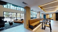 Located at 3560 Rambla Pl in Santa Clara, California, this single-floor community amenity space is a masterclass in contemporary design. Soaring double-height ceilings flood the interior with natural light through expansive floor-to-ceiling windows, creating an open and airy atmosphere throughout. Large-format porcelain tile flooring runs seamlessly across the space, grounding the various functional zones in a cohesive, clean aesthetic.

The lounge area is anchored by plush caramel-toned modular sofas and deep-hued accent chairs, all arranged around a bold vertical-slatted wood feature wall that frames a wall-mounted flat-screen television. Dramatic pendant lighting and exposed industrial ductwork add an urban edge to the otherwise refined interior.

A dedicated dining and collaboration zone features a long, built-in banquette with warm amber upholstery, complemented by sleek bistro-style tables and bar seating — ideal for both casual gatherings and focused work sessions. Natural wood-clad columns punctuate the space, adding warmth and visual structure.

The full-service kitchenette is outfitted with dark cabinetry, a white quartz countertop, stainless steel sink, built-in oven, and beverage refrigerator, all set against a striking geometric-patterned tile backsplash. Large glazed doors connect the interior directly to an outdoor patio, seamlessly blending indoor and outdoor living.
