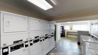 This shared laundry facility is designed to accommodate high-demand use, featuring a row of stacked commercial-grade washer and dryer units lining one wall. Each machine is equipped with a modern card payment interface, accepting major credit cards for convenient, cashless transactions. The units are finished in white and arranged in an orderly fashion, maximizing the available floor space.

The room transitions into an adjacent area visible through an open passageway, which houses a vending machine and a long utility table — ideal for folding and organizing laundry. A window in this secondary space allows natural light to filter in, brightening the otherwise functional environment.

The flooring throughout consists of a stone-patterned tile laid in a diagonal pattern, adding a subtle decorative element to the utilitarian space. Crown molding lines the ceiling, lending a slightly refined touch to the room. Overhead, a skylight panel provides additional illumination, keeping the main laundry area well-lit. A fire extinguisher is wall-mounted near the vending area, reflecting attention to safety standards. Overall, the facility is clean, organized, and practical.