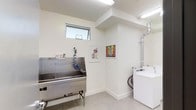 This well-appointed utility room serves a dual purpose, combining a dedicated pet grooming area with a functional laundry space. The room features clean white walls and light-toned tile flooring, creating a bright and hygienic environment. At the heart of the grooming area sits a stainless steel dog wash tub equipped with a mounted spray hose and accessory shelf, designed to make pet bathing efficient and convenient. A small horizontal window above the tub allows natural light to filter into the space. Fluorescent overhead lighting ensures consistent illumination throughout. On the opposite side of the room, full-sized washer and dryer units are neatly positioned alongside visible ventilation ductwork, underscoring the room's practical design. Colorful framed dog-themed artwork adorns the walls, adding a playful and welcoming character to an otherwise utilitarian space. The overall layout is thoughtfully organized, maximizing functionality while maintaining a clean, modern aesthetic suited for both pet care and household laundry needs.