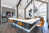 Located at 2789 Satellite Blvd in Duluth, GA, this single-story community clubhouse showcases a striking blend of modern and rustic design. The space is defined by dramatic vaulted ceilings with dark wood shiplap panels and exposed timber beam columns that anchor the open floor plan. Large floor-to-ceiling windows flood the interior with abundant natural light and offer views of the surrounding greenery, creating an airy, welcoming atmosphere throughout.

Wood-look plank flooring runs seamlessly across the entire space, complementing the warm tones of the amber and cognac upholstered seating. A long, sleek white quartz counter with bar-height seating serves as a central gathering point, while pendant lighting—ranging from cylindrical black fixtures to sculptural globe pendants—adds visual interest overhead.

The lounge area is furnished with comfortable seating arrangements ideal for relaxed conversation, while a dedicated media and beverage station features built-in cabinetry, a flat-screen TV, coffee service equipment, and a mini-refrigerator set against a textured tile accent wall. Rustic circular wood art pieces add a nature-inspired decorative touch nearby.

A game area with a full shuffleboard table provides recreational entertainment, and a glass-enclosed private room offers a more intimate setting for meetings or gatherings. The overall ambiance is sophisticated yet approachable, balancing contemporary finishes with warm, natural design elements.