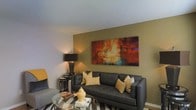 This single-floor space presents a well-appointed living area designed with a contemporary aesthetic and a warm, inviting ambiance. The living room is anchored by a sleek, dark leather sofa accented with gold and beige throw pillows, complementing the earthy olive-toned accent wall that serves as a striking visual focal point. A large, vibrant abstract canvas painting in rich shades of burnt orange, red, and teal commands attention above the sofa, adding an artistic and dynamic energy to the room. A round glass-top coffee table sits at the center, contributing to the open, airy feel of the space while displaying tasteful decorative objects. Ambient lighting is provided by a tall, modern table lamp with a dark shade, casting a warm glow throughout the room. The neutral ceiling and light-colored trim create a clean contrast against the deeper wall tones, enhancing the overall sense of depth and dimension. The flooring features a patterned area rug that adds texture and visual interest underfoot. Together, the carefully curated furnishings and color palette create a cohesive, sophisticated living environment that balances comfort with style.