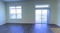 This single-floor space offers a clean, modern aesthetic defined by its light-toned walls and rich dark hardwood flooring that runs throughout the main living area. Natural light is a defining feature, streaming in generously through a large multi-pane window on one side and a full sliding glass door on the other, creating an open, airy atmosphere throughout the day. The sliding glass door leads directly to an outdoor balcony enclosed by a white railing, offering views of the surrounding neighborhood and beyond. The transom window above the sliding door further enhances the brightness of the space, drawing in additional light from above. The interior presents a neutral, versatile palette with crisp white trim and baseboards that contrast cleanly against the warm-toned flooring. The layout is open and unobstructed, allowing for flexible furniture arrangement and a spacious feel. The combination of abundant glazing, elevated balcony access, and a well-maintained interior makes this a light-filled, functional space suited to a variety of uses.