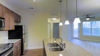Located at 320 The Greens Cir in Raleigh, NC, this single-floor residence offers a thoughtfully designed layout with stylish finishes throughout. The kitchen serves as a central highlight, showcasing granite countertops with an undermount double-basin stainless steel sink, dark cabinetry, and a stainless steel refrigerator. Elegant pendant lighting hangs above the kitchen island, complementing the soaring ceiling height that gives the space an open, airy feel. Natural light filters in through a window, brightening the area and enhancing the warmth of the hardwood-style flooring that runs throughout. Crisp white walls and clean architectural lines contribute to a modern, welcoming ambiance. A pass-through hallway connects the kitchen to the additional rooms, offering an efficient and cohesive flow across the floor plan. The high ceilings, quality countertop materials, and contemporary lighting fixtures all work together to create a polished interior that balances practicality with refined design. This Raleigh residence presents a comfortable and well-appointed living environment suitable for everyday living.