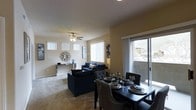 This single-story space features an open-plan design that seamlessly integrates the dining and living areas into one cohesive environment. The living room is anchored by plush carpeting and a ceiling fan with integrated lighting, creating a comfortable and functional atmosphere. Large windows fitted with vertical blinds allow generous amounts of natural light to fill the space, contributing to a bright and airy feel. A sliding glass door provides direct access to an outdoor area, further enhancing the sense of openness and connectivity to the exterior.

The dining area is positioned adjacent to the living room, defined by a dark-toned dining table with tufted upholstered chairs, offering a stylish setting for meals. The living room features a coordinated dark blue sofa set arranged around a central area, complemented by a console table along the wall. Neutral-toned walls and carpeting provide a versatile backdrop that complements the furnished interior. The overall layout is efficient and well-proportioned, making practical use of the available space while maintaining a clean, modern aesthetic throughout.