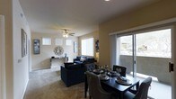 This single-story space features an open-plan design that seamlessly integrates the dining and living areas into one cohesive environment. The living room is anchored by plush carpeting and a ceiling fan with integrated lighting, creating a comfortable and functional atmosphere. Large windows fitted with vertical blinds allow generous amounts of natural light to fill the space, contributing to a bright and airy feel. A sliding glass door provides direct access to an outdoor area, further enhancing the sense of openness and connectivity to the exterior.

The dining area is positioned adjacent to the living room, defined by a dark-toned dining table with tufted upholstered chairs, offering a stylish setting for meals. The living room features a coordinated dark blue sofa set arranged around a central area, complemented by a console table along the wall. Neutral-toned walls and carpeting provide a versatile backdrop that complements the furnished interior. The overall layout is efficient and well-proportioned, making practical use of the available space while maintaining a clean, modern aesthetic throughout.