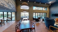 This two-story amenity space is designed to serve as a dynamic social and work hub, blending modern aesthetics with functional versatility. The main gathering area commands immediate attention with its soaring double-height ceilings, dramatic pendant lighting, and expansive floor-to-ceiling windows and sliding glass doors that open to views of a resort-style pool courtyard. Natural light floods the hardwood floors, creating a bright and welcoming atmosphere throughout.

The fully equipped kitchen features stainless steel appliances, dark cabinetry with tile backsplash, a large island with bar seating, and direct pool access via glass doors—ideal for events and casual gatherings. Adjacent co-working and lounge areas offer flexible seating arrangements beneath pendant lights, with moss-art wall installations and a large display screen providing a creative, productive environment.

A dedicated media lounge includes a dark, intimate atmosphere with sectional leather seating, a glass coffee table, and built-in cabinetry. A second sitting area on the lower level showcases a striking geometric feature wall, mid-century modern furnishings, and an upper-level mezzanine visible above. Throughout, the design palette of blue-toned ceilings, warm wood floors, and curated décor establishes a cohesive, contemporary ambiance.
