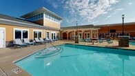 Located at 700 Berkeley Place Circle in Clemson, South Carolina, this outdoor amenity area centers around a generously sized, resort-style swimming pool with clear turquoise water. The pool features a curved, freeform design with multiple entry points, including wide shallow-entry steps that provide easy access from the surrounding concrete deck. Depth markers are visible along the pool's edge, indicating varied water depths suited for a range of swimmers.

The expansive pool deck is lined with rows of blue and white lounge chairs, providing ample space for sunbathing and relaxation. Decorative stone pillar accents are positioned throughout the deck, adding a refined architectural detail to the outdoor setting. Shade sail structures are installed in a corner of the deck area, offering relief from direct sun exposure.

Adjacent to the pool, a well-maintained clubhouse building features warm yellow and orange exterior siding with a covered porch area outfitted with casual seating. The structure includes restroom and facility access, and its two-story section features large windows overlooking the pool. A classic lamppost stands prominently near the pool's edge, enhancing the community aesthetic.

The entire area is enclosed by an iron fence and bordered by mature pine and deciduous trees, providing a natural, private backdrop. The space is open to the sky, making it an ideal venue for outdoor recreation in a well-designed community setting.