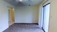 This single-floor residential space offers a straightforward and functional layout designed for comfortable everyday living. The main living area is finished with wall-to-wall carpeting and features light, neutral-toned walls that create a calm and versatile backdrop suitable for a variety of interior styles. A ceiling fan is centrally mounted overhead, providing both air circulation and a practical lighting source for the room. A large sliding glass door along one wall allows for an abundance of natural light to fill the space, while vertical blinds offer adjustable privacy and light control. The door also provides direct access to an exterior area, adding convenience and connectivity to the outdoors. White baseboards run cleanly along the perimeter of the room, adding a crisp contrast to the soft wall tones. Multiple electrical outlets are positioned along the walls, offering practical utility throughout the space. A doorway visible from the living area leads further into the interior of the home, suggesting additional rooms beyond. The overall layout is clean and open, providing a blank canvas well-suited for personalization and customization to meet a variety of living needs.