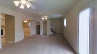 This two-story home offers a practical and open layout designed for comfortable living. The main level features a spacious living area finished with light-toned wall-to-wall carpeting that extends throughout, creating a cohesive and neutral foundation. Ceiling fans are installed throughout the main floor, including in the primary living space and an adjoining room visible through an open doorway, ensuring good air circulation and comfort year-round.

The living area flows naturally toward a staircase fitted with a classic wrought-iron railing, providing access to the second floor. Nearby, a set of double doors hints at additional storage or closet space, adding functional utility to the layout. A small window on the far wall allows a modest amount of natural light to filter into the space.

An adjacent room, partially visible through an open doorway, features its own ceiling fan and a door to the exterior, suggesting it may serve as a secondary living or dining area. The overall aesthetic is clean and understated, with neutral beige walls and minimal ornamentation, offering a versatile blank canvas adaptable to a variety of interior styles.