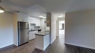 This well-appointed single-floor residence offers a clean, contemporary aesthetic throughout its thoughtfully designed layout. The kitchen is a standout feature, showcasing crisp white cabinetry that extends to the ceiling, dark contrasting countertops, and stainless steel appliances including a full-sized refrigerator. A peninsula island provides additional prep space and serves as a natural divider between the kitchen and the adjoining areas. The sink is positioned beneath a flush-mounted fluorescent light, ensuring ample task lighting. Dark hardwood flooring runs consistently throughout the space, grounding the interior with warmth and contrast against the light grey walls and white trim. The hallway leading from the kitchen connects seamlessly to the rest of the unit, with a glimpse of a bathroom visible at the end, featuring white finishes consistent with the overall design language. The neutral grey wall tones and white baseboards create a cohesive, modern palette that is both versatile and inviting. Flush-mount ceiling fixtures provide even ambient lighting throughout. The overall ambiance is bright, clean, and move-in ready, reflecting a practical yet stylish approach to residential design.