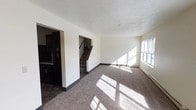 This two-story residence offers a straightforward and functional layout designed for comfortable everyday living. The main level features an open living area finished with neutral carpet flooring that transitions into tile near the kitchen entry, creating a clear distinction between living and functional spaces. Large windows on the main floor allow generous amounts of natural light to fill the room, casting bright patterns across the floor and contributing to an airy, welcoming atmosphere. The textured ceiling adds a classic touch throughout the space. Dark-finished cabinetry is visible in the kitchen area, providing a strong contrast against the light-toned walls and lending a modern accent to the overall interior palette. A carpeted staircase, framed by a clean archway opening, connects the main level to the upper floor, offering a seamless flow between the two stories. The baseboard heating units along the walls are a practical feature suited for year-round comfort. The neutral color scheme of white walls and understated finishes throughout creates a versatile backdrop adaptable to a variety of interior styles and furnishings. Overall, the space presents a clean, functional environment with a balance of natural light and practical design across both levels.