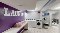 This single-floor amenity space is divided into two distinct functional areas: a laundry room and a bicycle storage room. The laundry room is visually distinctive, featuring a bold purple accent wall with large white "LAUNDRY" lettering that creates a lively focal point. The room is equipped with multiple stacked and front-loading washers and dryers, a digital payment terminal, informational boards, and a built-in folding/seating bench for added convenience. Light gray walls, white tile flooring, and recessed ceiling lighting contribute to a clean, bright atmosphere. A stainless steel waste bin and a fire alarm are also present, reflecting attention to safety and cleanliness. The adjacent bicycle storage room offers a generous open floor plan with a speckled concrete floor and a suspended drop ceiling with flush-panel lighting. Multiple wall-mounted and freestanding bike racks line the perimeter, providing organized and secure storage for residents' bicycles. The neutral gray walls and clean layout keep the space practical and easy to navigate. A entrance mat bearing the building name "Roosevelt Apartments" connects the two areas, indicating this facility serves a residential building. Together, these two rooms form a well-appointed, functional amenity area designed to meet everyday resident needs.