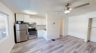 This single-floor space offers a practical and modern layout centered around an open-concept kitchen and living area. The kitchen is well-equipped with stainless steel appliances, including a refrigerator, electric range, and microwave, all complemented by crisp white cabinetry and a subtle mosaic tile backsplash that adds a touch of texture and visual interest. Light-toned countertops and a stainless steel sink complete the functional cooking space. The kitchen flows seamlessly into the living area, which features light hardwood-style flooring that runs throughout, giving the space a cohesive and airy feel. A ceiling fan with an integrated light fixture is centrally positioned in the living area, providing both airflow and ambient lighting. The textured popcorn ceiling adds a classic finish overhead. A doorway visible from the main space leads to an additional area, suggesting a separate room or hallway beyond. The overall palette is clean and neutral, with white walls and light flooring creating a bright, welcoming environment. The open layout and functional design make this a straightforward, move-in-ready space well-suited for everyday living.
