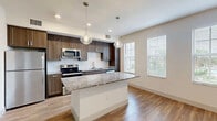 Located at 3200 Champion Ring Rd in Fort Myers, Florida, this one-bedroom, one-bathroom residence offers a thoughtfully designed single-floor layout. The open-concept living area seamlessly connects the kitchen, dining, and living spaces, creating an inviting and functional environment. The kitchen is a standout feature, showcasing rich dark wood cabinetry, granite countertops, a white subway tile backsplash, and a full suite of stainless steel appliances including a refrigerator, range, and built-in microwave. A central island with a granite surface and elegant pendant lighting serves as both a prep area and casual dining spot. Large windows allow natural light to fill the space, offering views of the surrounding greenery. Wood-look flooring runs throughout the unit, adding warmth and continuity. The bedroom is a calm, neutral retreat with ample wall space and a closet with bifold doors, complemented by additional built-in shelving for extra storage. The bathroom features large-format marble-style tile surrounding a soaking tub and shower combination, paired with matching granite countertops. A dedicated laundry area and hallway complete this efficiently planned home.