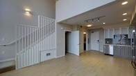 This well-designed two-story loft-style space offers a thoughtfully open layout that immediately conveys a sense of height and airiness. The main level features light-toned hardwood flooring that runs throughout, lending warmth and continuity to the space. Soaring double-height ceilings define the living area, drawing the eye upward and creating an expansive, loft-like atmosphere rarely found in comparable spaces.

A clean white staircase with vertical baluster railings rises prominently along one wall, connecting the two levels while serving as an architectural focal point. The upper level overlooks the main floor through an open railing, reinforcing the sense of vertical space and connectivity between floors.

Track lighting mounted on the ceiling provides targeted, adjustable illumination across the main living area, complementing the recessed lighting found throughout. The entryway is clearly defined near the front door, which features a modern keypad entry system for added convenience and security.

Just off the main living area, open doorways lead to additional rooms including what appears to be a closet and a bathroom or utility space, offering practical functionality. A partial view of a kitchen area is visible to one side, featuring white cabinetry and modern appliances. The neutral color palette of warm beige walls and white trim throughout creates a clean, inviting, and versatile environment.