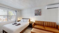 Located at 425 Second St N in the heart of downtown Saint Petersburg, Florida, this single-floor studio unit offers a thoughtfully designed layout that maximizes functionality within a compact footprint. The main living area is finished with light-toned laminate wood flooring and bright white walls accented by a textured tile panel behind the king-sized bed, lending a clean and modern aesthetic throughout. A mid-century-style caramel leather sofa, wooden coffee table, and wall-mounted flat-screen TV create a comfortable living zone that flows seamlessly alongside the sleeping area. Natural light enters through a window fitted with blinds near the entry door, keeping the space feeling open and airy. A wall-mounted split A/C unit ensures climate control year-round. The compact kitchenette features white cabinetry, a mosaic tile backsplash, an electric range, refrigerator, microwave, and a coffee maker — providing all essentials for self-sufficient living. A dedicated walk-in closet offers open shelving and hanging space, adding practical storage. The bathroom is finished with large-format white tile flooring and marble-look wall tile surrounding the tub-shower combination, creating a clean and polished feel. A small dining area with a round table and two chairs completes the studio's versatile layout.