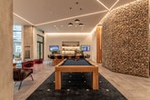 This contemporary amenity space is designed to offer a dynamic and social environment across a single open floor. The entry is announced by a striking natural wood-paneled feature wall with inset lighting strips, complemented by deep navy blue accent walls that establish a bold visual identity throughout. Large floor-to-ceiling windows allow generous natural light to fill the space, with views of the surrounding streetscape visible from the glass-fronted lobby area, which features sleek stone tile flooring and linear LED ceiling details.

The main lounge area is anchored by a solid wood pool table set atop a dark area rug, positioned beneath geometric LED light fixtures that trace angular patterns across the ceiling. Adjacent to the game area, a fully equipped bar-height counter with dark stone tops and warm wood cabinetry provides ample seating for socializing, flanked by a distinctive gabion stone column that adds a raw, textural contrast to the polished interior.

A dedicated lounge nook offers a comfortable sectional sofa in rich cognac leather, layered with colorful throw pillows and paired with a vibrant striped area rug. A wall-mounted large-screen TV, wooden media console, and curated artwork complete the entertainment zone. Throughout, the design balances warm natural materials with modern finishes to create an engaging, well-appointed communal space.